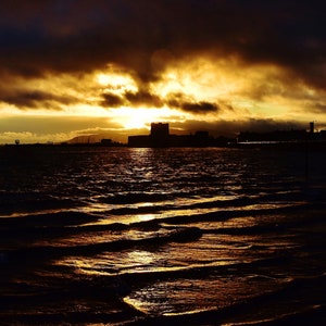 May include: A silhouette of a city skyline with a dark, cloudy sky behind it. The sun is setting behind the city, casting a golden glow on the clouds. The water in the foreground is dark and choppy.