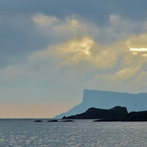 Può includere: Una vista panoramica di una costa rocciosa con una scogliera drammatica in lontananza. Il cielo è di un azzurro tenue con nuvole bianche soffici e un raggio di sole che filtra attraverso.