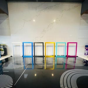 May include: Six colorful picture frames in white, blue, black, yellow, turquoise, and pink are arranged on a black stovetop. The frames are rectangular with a simple, modern design. The stovetop has a reflective surface.