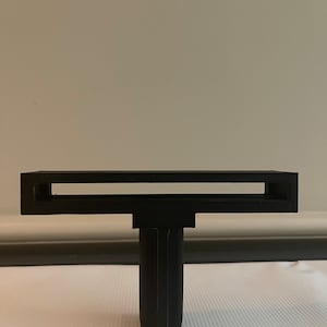 Può includere: Un oggetto di plastica nera a forma di T con un'apertura rettangolare nella parte superiore. L'oggetto è progettato per contenere un piccolo oggetto, come un telefono o un tablet.