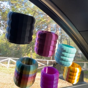 May include: Six colourful, striped, cylindrical car air fresheners hanging from the rear-view mirror of a car. The air fresheners are in shades of purple, blue, yellow, green, and black.