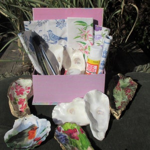 May include: A pink and white striped box filled with craft supplies for a DIY project. The box contains fabric scraps, paintbrushes, a bottle of Mod Podge, and oyster shells. The shells are decorated with floral patterns and a bird.