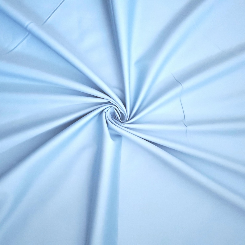May include: A pale blue fabric is arranged in a spiral, with folds and shadows creating depth. The fabric has a smooth texture and a slight sheen. The colour is a soft, pastel blue, and the fabric appears to be lightweight.