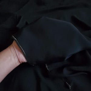 Op de afbeelding: Een effen zwarte stof met een gladde, matte textuur en een subtiele glans. Het materiaal is voorzien van een delicaat, dun zilverkleurig metallic draadje dat langs de afgewerkte rand is gestikt, wat een verfijnd contrast vormt met de stof.
