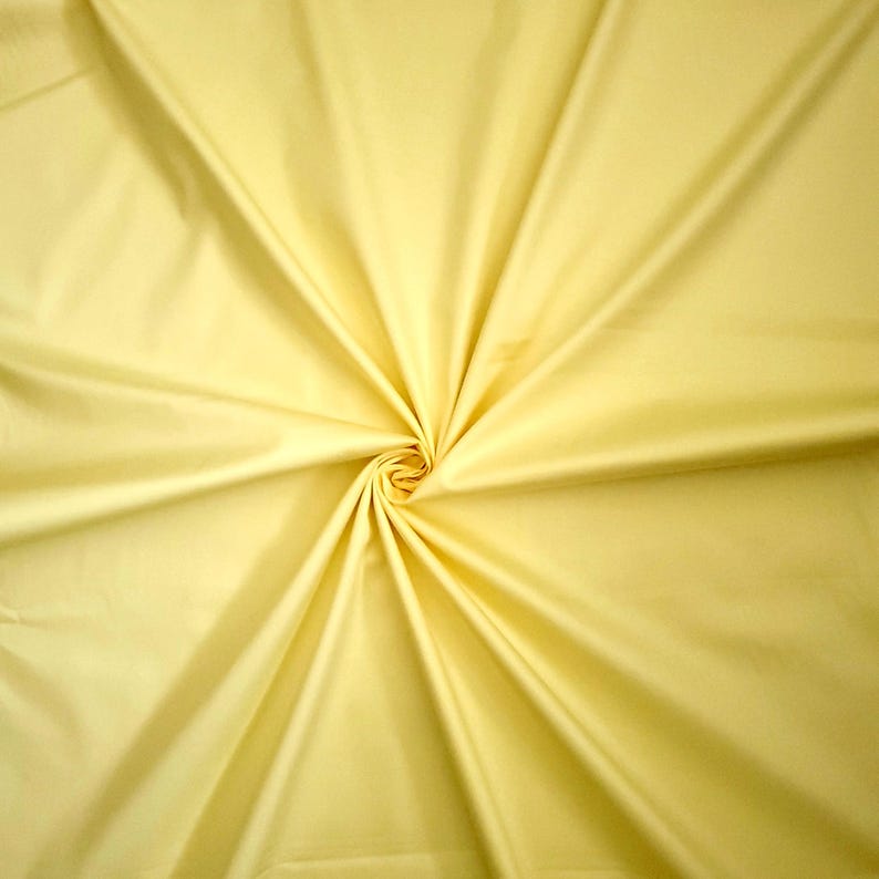 May include: A close-up of pale yellow fabric, tightly swirled at the centre. Folds radiate outwards, creating a starburst effect. The material appears smooth and possibly silky, with subtle tonal variations. The fabric is likely for clothing or home decor.