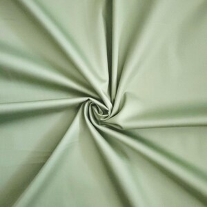 May include: A close-up of a sage green fabric with a smooth, matte finish. The fabric is gathered and swirled in the centre, creating a spiral pattern. The folds and shadows add depth and texture to the material.