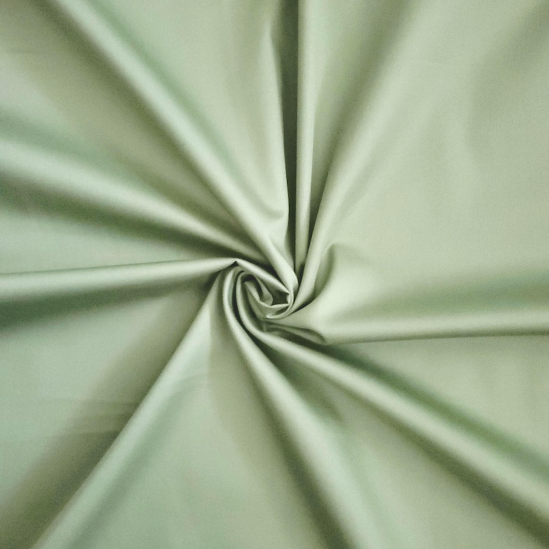 May include: A close-up of a sage green fabric with a smooth, matte finish. The fabric is gathered and swirled in the centre, creating a spiral pattern. The folds and shadows add depth and texture to the material.