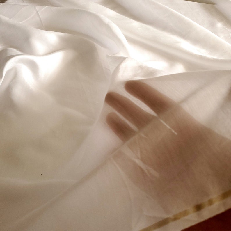Op de afbeelding: Een stuk witte stof met een lichtgewicht, semi-transparante textuur die in zachte plooien valt. Het materiaal is dun genoeg om het silhouet van vingers door het oppervlak heen zichtbaar te maken, wat de doorschijnende kwaliteit benadrukt.