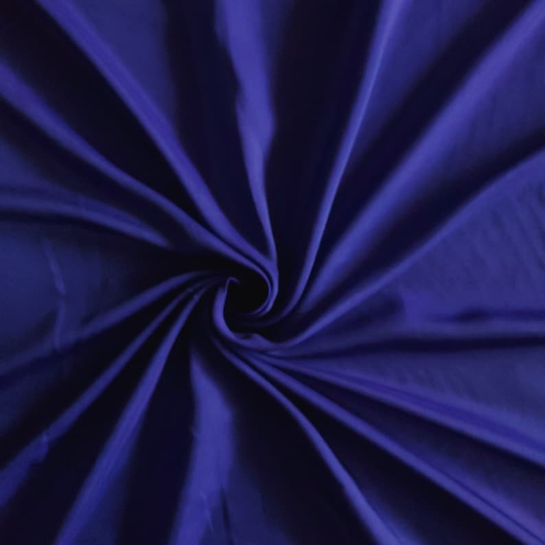 Op de afbeelding: Een diep koningsblauwe stof met een glad, licht reflecterend oppervlak. Het materiaal is in een centrale spiraal bijeengebracht, waardoor stralende plooien en schaduwen ontstaan die de textuur en de rijke, verzadigde blauwe kleur benadrukken.