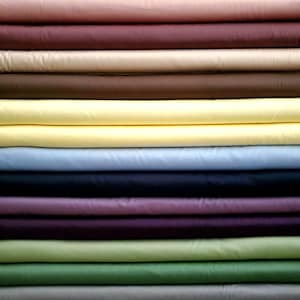 May include: A pile of folded fabric in a range of colours: pink, burgundy, beige, brown, yellow, light blue, navy, purple, green, and grey. The fabric looks smooth, possibly cotton or a similar material.