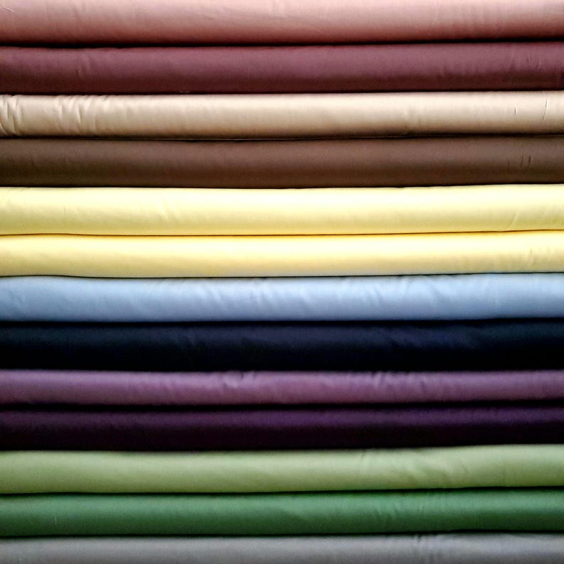 May include: A pile of folded fabric in a range of colours: pink, burgundy, beige, brown, yellow, light blue, navy, purple, green, and grey. The fabric looks smooth, possibly cotton or a similar material.