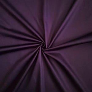 May include: A close-up of a deep purple fabric with a swirling design. The material has a smooth, satin-like texture, gathered at the centre, creating radiating folds and shadows. The colour is a rich, dark shade of purple.