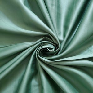 May include: A close-up shot of sage green fabric, showing a swirling pattern of folds and creases. The fabric has a smooth, lustrous finish, reflecting light and creating highlights and shadows. The material appears to be lightweight, possibly silk or satin, with a soft, flowing drape.
