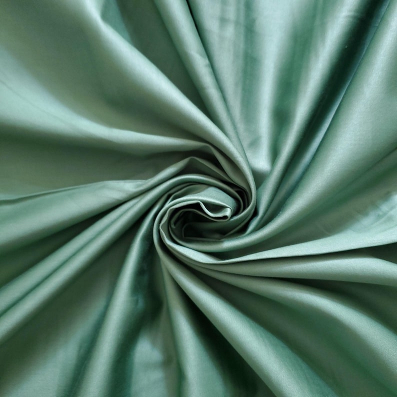 May include: A close-up shot of sage green fabric, showing a swirling pattern of folds and creases. The fabric has a smooth, lustrous finish, reflecting light and creating highlights and shadows. The material appears to be lightweight, possibly silk or satin, with a soft, flowing drape.