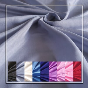 Op de afbeelding: Een lichtblauwe stof met een gladde, matte textuur en zachte plooien. Onder de hoofdstof toont een horizontale strook acht kleurvariaties van hetzelfde materiaal: zwart, lichtblauw, gebroken wit, blauw, donkerblauw, paars, roze en rood.