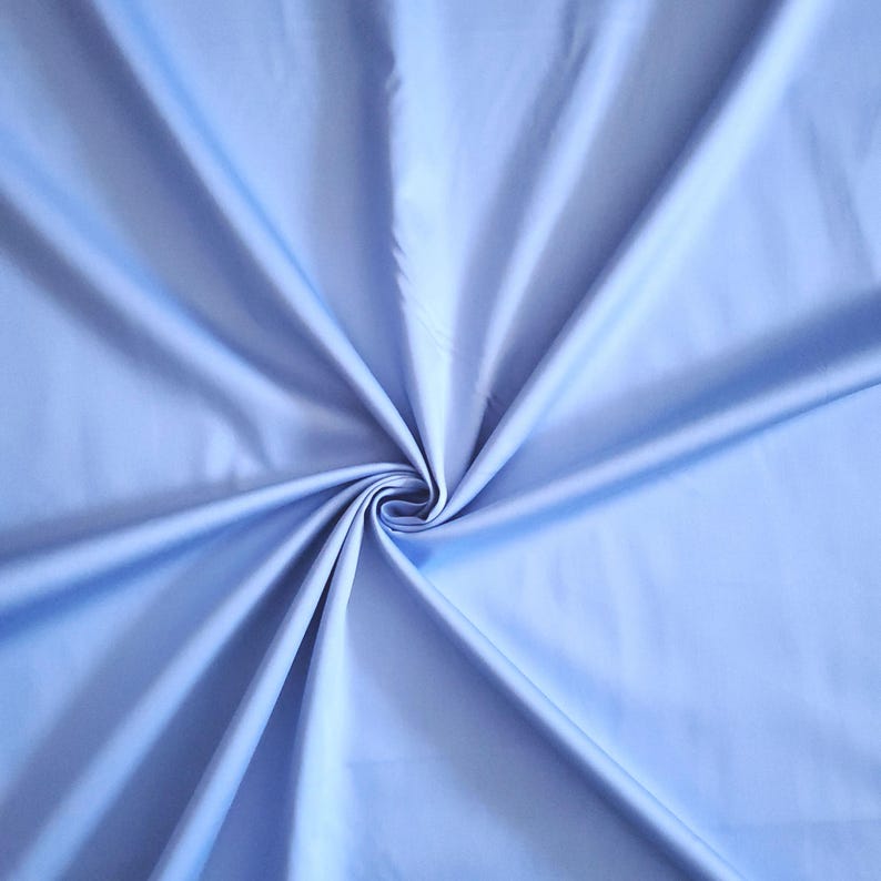 May include: A close-up of a light blue fabric with a smooth, silky texture. The fabric is gathered in a spiral pattern, creating soft folds and shadows. The colour is a pale, calming shade of blue.