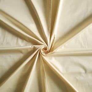May include: A close-up of a cream-coloured fabric with a smooth, shiny finish. The material is arranged in a spiral, with folds and shadows adding depth and texture. The fabric looks lightweight and flowing, suitable for clothing or home furnishings.