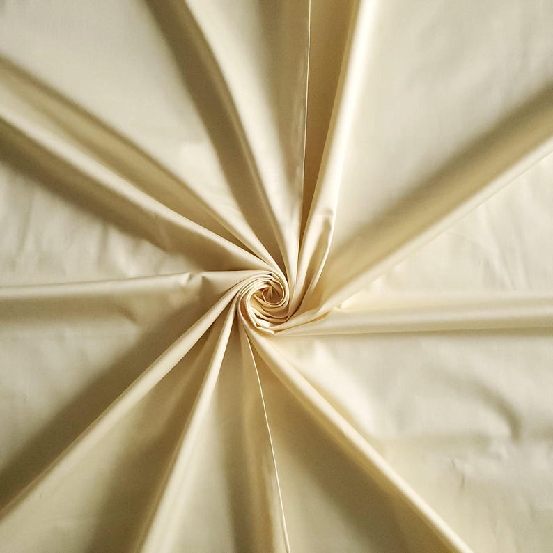 May include: A close-up of a cream-coloured fabric with a smooth, shiny finish. The material is arranged in a spiral, with folds and shadows adding depth and texture. The fabric looks lightweight and flowing, suitable for clothing or home furnishings.