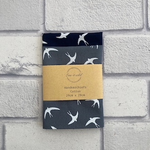 May include: Three cotton handkerchiefs in shades of blue, stacked and wrapped with a brown paper band. Each handkerchief features a white swallow bird pattern. The paper band reads "Sew it wild" and "Handkerchiefs Cotton 29cm x 29cm."