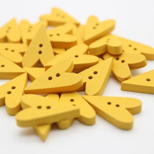 Könnte beinhalten: Ein Haufen gelber, herzförmiger Holzknöpfe mit jeweils zwei Löchern. Die Knöpfe sind in einem zufälligen Muster angeordnet.