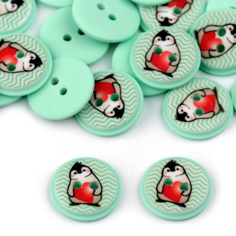 May include: A pile of mint green buttons with a penguin holding a red heart design. The buttons have two holes.
