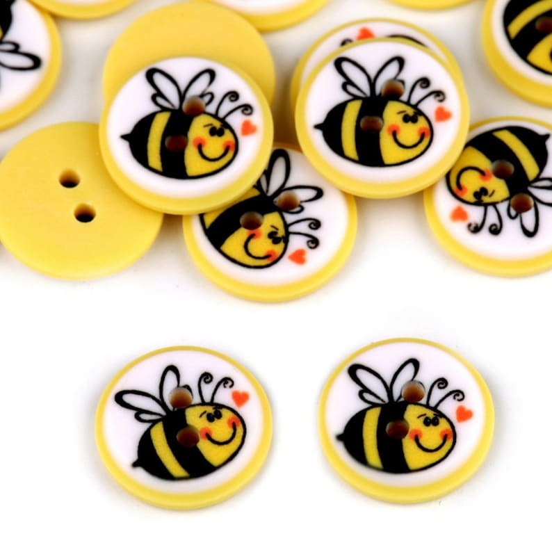 May include: A collection of yellow plastic buttons with a cartoon bee design. The bees are black and yellow with white wings and have a smiling face with two red hearts.
