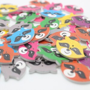 Könnte beinhalten: Ein Haufen bunter Holz-Knöpfe mit Waschbär-Gesichtern. Die Knöpfe sind in verschiedenen Farben, darunter Rot, Blau, Grün, Gelb und Lila. Jeder Knopf hat ein schwarzes Waschbär-Gesicht mit weißen Augen und einer schwarzen Nase.