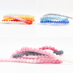 Könnte beinhalten: Sortiment an bunten Pompon-Borten in verschiedenen Farben, darunter Rosa, Weiß, Grau, Blau und Orange. Die Borte ist aus weichem, flauschigem Material gefertigt und perfekt, um jedem Projekt einen Hauch von Verspieltheit zu verleihen.