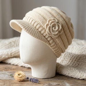 May include: Cream-colored knit hat with a small brim and a decorative flower on the side. The hat is displayed on a mannequin head. The knit pattern is visible, and the hat appears soft and warm. A cozy accessory for cooler weather.