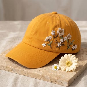 May include: Mustard yellow baseball cap with embroidered white and green floral design. The cap features a curved brim and a top button. A daisy and a gerbera daisy are next to the cap.