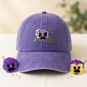 Puede incluir: Gorra de béisbol morada con un diseño de bordado de flores de pensamiento. La flor presenta pétalos morados, negros y amarillos con hojas verdes. Dos pensamientos reales se colocan cerca de la gorra.