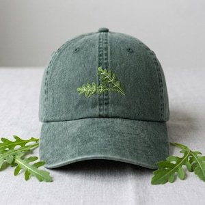 May include: A green baseball cap with a curved peak and a button on top. The cap has a light green embroidered fern design on the front. The cap is made of a textured fabric and is displayed on a neutral surface.
