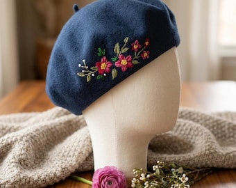Op de afbeelding: Een marineblauwe baret met geborduurde bloemen in roze, rood en groen. De baret wordt getoond op een mannequinhoofd. Een roze bloem en takjes witte bloemen staan op de voorgrond.