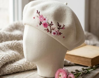 May include: Cream-colored beret with embroidered pink and white floral design. The beret is displayed on a mannequin head. The flowers are clustered on the side of the hat, adding a touch of elegance. The beret is made of a soft, textured material.