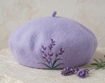 Può includere: Un basco lilla con un motivo di spighe di lavanda ricamate. Il basco è realizzato in un materiale morbido, simile al feltro. Un rametto di lavanda è posto accanto al basco.