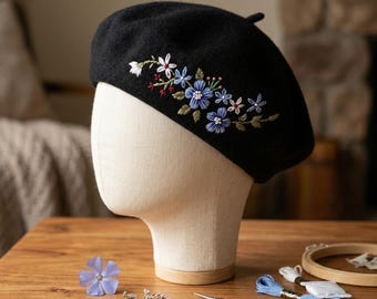 May include: A black beret with embroidered floral design in shades of blue, white, and red. The beret is displayed on a mannequin head. Embroidery supplies, including scissors and thread, are on the wooden surface.
