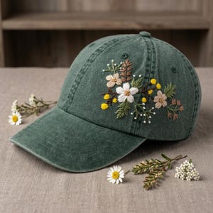 May include: A green baseball cap with an embroidered floral design. The embroidery features white, yellow, and tan flowers with green leaves. The cap is made of a soft, worn fabric.