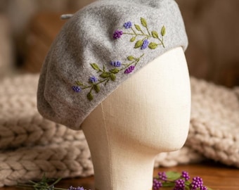 May include: A gray wool beret with embroidered floral details. The beret features a delicate design of purple and blue flowers with green leaves. The hat is displayed on a mannequin head, showcasing its shape and style. A sprig of lavender is visible.