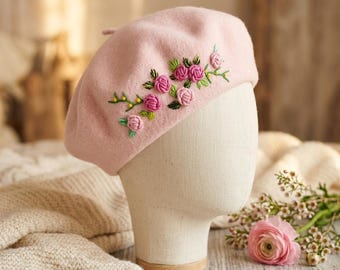May include: A light pink beret with embroidered pink roses and green leaves. The beret is displayed on a mannequin head. The beret is a stylish accessory, perfect for adding a touch of elegance to any outfit. The beret is made of a soft, felt-like material.