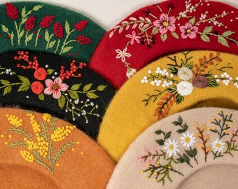 Puede incluir: Cinco boinas de fieltro en diferentes colores, cada una con un diseño floral bordado único. Las boinas están apiladas una encima de la otra, con la boina verde en la parte superior y la boina beige en la parte inferior.