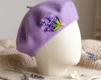 May include: A lavender-colored beret with a small, decorative cluster of purple flowers and green leaves. The beret is displayed on a mannequin head. The hat is made of a soft, felt-like material.