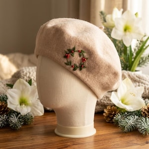May include: A beige beret with an embroidered wreath of green leaves and red berries. The hat is displayed on a mannequin head. The beret is made of a soft, wool-like material. The background includes white flowers and pine cones.