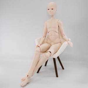 Pode incluir: Uma boneca articulada de cor pálida com um acabamento mate suave, sentada numa pequena cadeira branca com pernas castanho-escuras. A boneca apresenta uma expressão facial neutra, musculatura definida no tronco e articulações visíveis nos ombros, cotovelos, pulsos, ancas, joelhos e tornozelos, permitindo uma postura sentada natural.