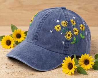 May include: A denim blue baseball cap with embroidered yellow sunflowers and white star accents. The cap features a curved brim and a classic six-panel design. The sunflowers are detailed with green leaves and brown centers.