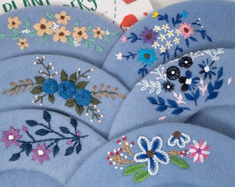 Op de afbeelding: Vijf blauwe wollen baretten met verschillende geborduurde bloemenmotieven. De motieven omvatten gele, witte, blauwe, roze en groene bloemen.