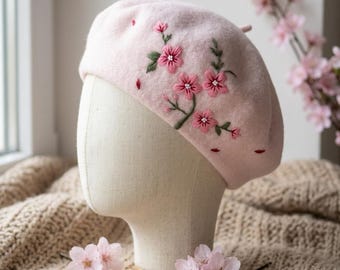 May include: A light pink beret with embroidered pink cherry blossoms and green leaves. The beret is displayed on a mannequin head. The hat is made of a soft material and is a stylish accessory.
