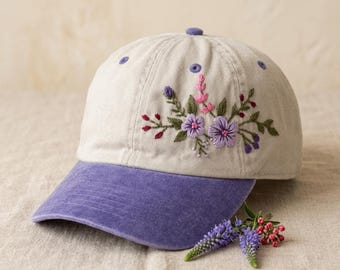 Può includere: Cappellino da baseball beige con visiera e bottone viola. Il cappellino è ricamato con un motivo floreale nei toni del viola, rosa e verde. Un rametto di fiori viola e bacche rosa è appoggiato vicino.