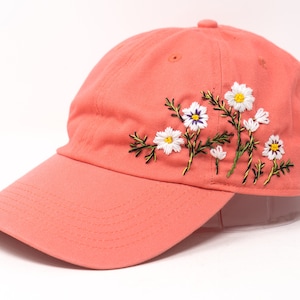 May include: A coral pink baseball cap with embroidered white daisies and green stems.