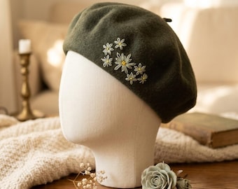May include: An olive green beret with embroidered white and yellow daisy flowers. The beret is displayed on a mannequin head. The background includes a wooden table, a candle holder, and a book.