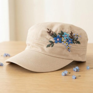 May include: A beige military-style cap featuring an embroidered floral design on the side. The embroidery consists of blue flowers, green leaves, and small yellow accents. The cap has a structured crown and a stiff, curved brim in a solid beige fabric.